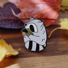 Chubby gray raccoon enamel pin holding a giant acorn with both arms, featuring an oak leaf and fall colors. Designed by Noristudio.