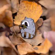 Chubby gray raccoon enamel pin holding a giant acorn with both arms, featuring an oak leaf and fall colors. Designed by Noristudio.