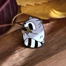 Chubby gray raccoon enamel pin holding a giant acorn with both arms, featuring an oak leaf and fall colors. Designed by Noristudio.
