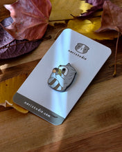 Chubby gray raccoon enamel pin holding a giant acorn with both arms, featuring an oak leaf and fall colors. Designed by Noristudio.