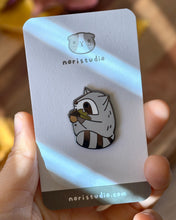 Chubby gray raccoon enamel pin holding a giant acorn with both arms, featuring an oak leaf and fall colors. Designed by Noristudio.